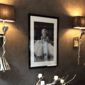 Ein Bild mit einer schwarz-weißen Fotografie einer Person in einem Ballkleid hängt zwischen zwei Wandleuchten an einer dunklen Wand.