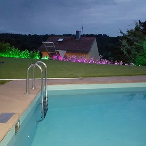 Ein Pool mit blauem Wasser und einer Treppe im Vordergrund, umgeben von beleuchtetem Gras und einem Haus im Hintergrund bei Nacht.