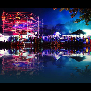 Eine beleuchtete Freiluft-Veranstaltung mit Menschen, Zelten und einer beleuchteten Struktur, die sich spiegelnd im Wasser reflektiert.