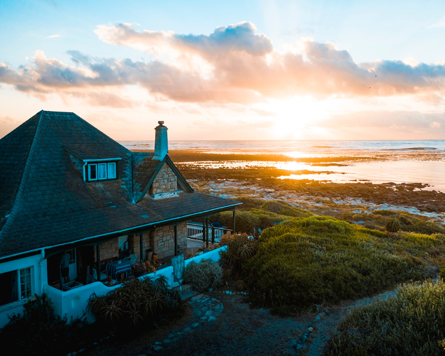 Ein Haus mit Schieferdach steht am Meer bei Sonnenuntergang, umgeben von Grünflächen und einem ruhigen Horizont.