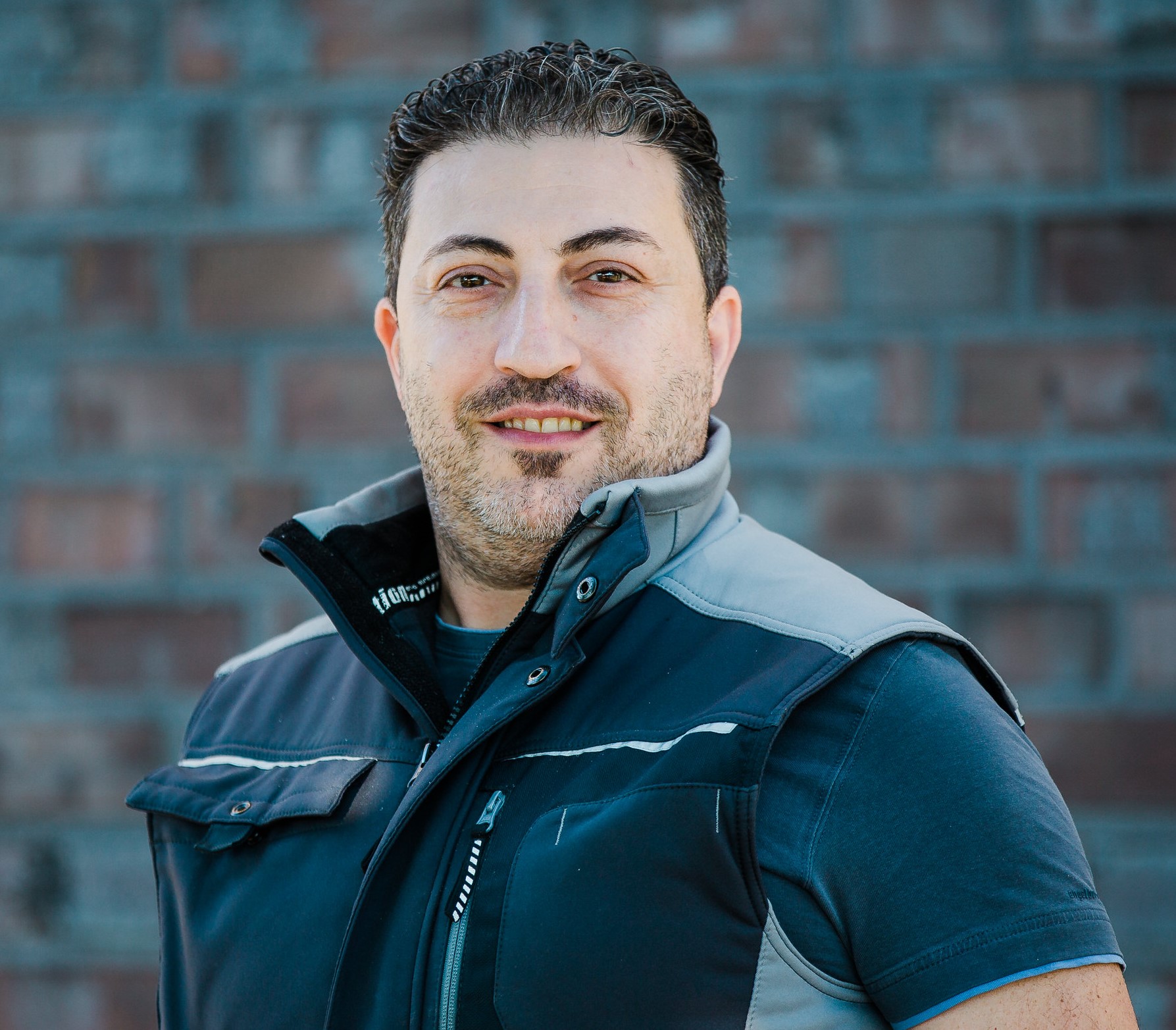 Ein Mann mit Bart und kurzen Haaren trägt eine Jacke und lächelt vor einem Backsteinhintergrund.
